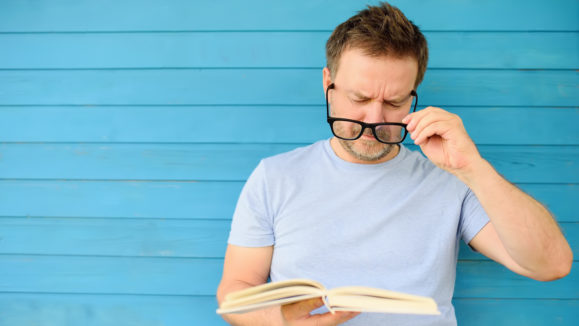 Portrait of mature man with big black eye glasses trying to read book but having difficulties seeing text because of vision problems